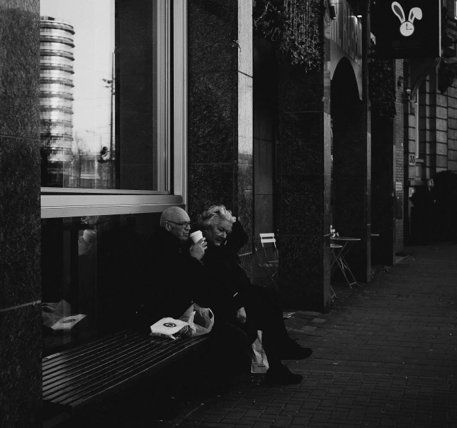 a black and white photo of a man sitting on a bench, a black and white photo, by Tamas Galambos, pexels contest winner, people waiting in bus stop, having a snack, couple, flowers around