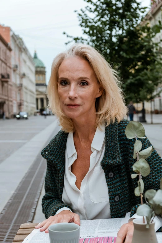 a woman sitting at a table with a cup of coffee, a portrait, inspired by Ulrika Pasch, pexels contest winner, wearing green jacket, standing in street, cardigan, magdalena andersson