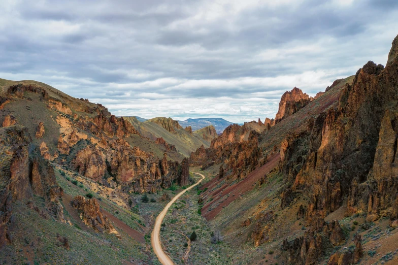 a dirt road winding through a canyon on a cloudy day, by Jeffrey Smith, unsplash contest winner, art nouveau, red narrow lava rivers, majestic spires, oregon trail, wide high angle view