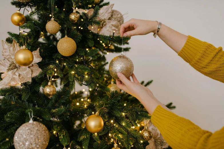 a woman decorating a christmas tree with gold ornaments, by Julia Pishtar, pexels, semi-realistic, thumbnail, high quality image, golden orbs