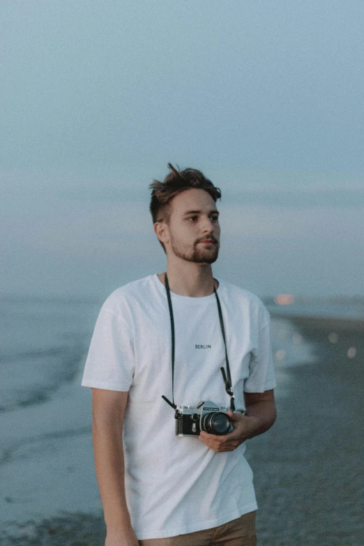 a man standing on a beach holding a camera, avatar image, jovana rikalo, dressed in a white t-shirt, professional profile picture