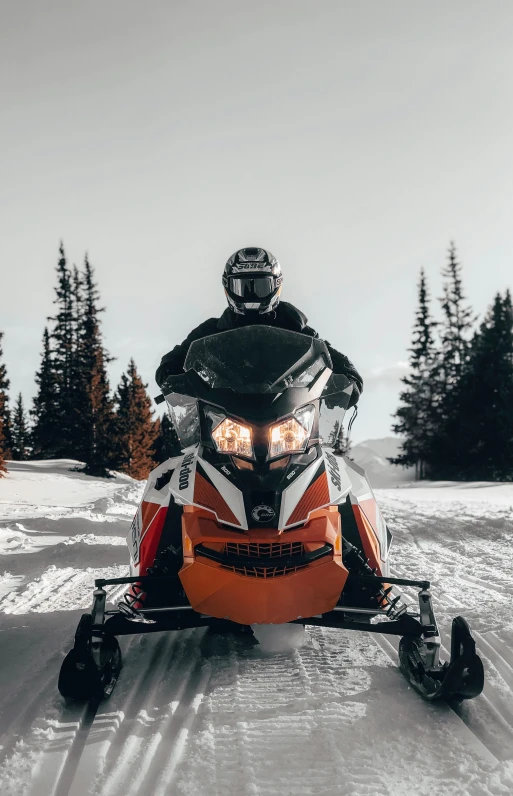 a person riding a snowmobile in the snow, front facing the camera, orange and white color scheme, dark setting, uploaded