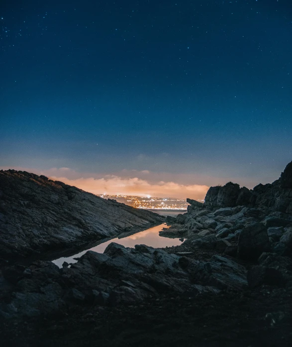 a body of water surrounded by rocks under a night sky, by Sebastian Spreng, high-quality photo, distant town lights, full frame image