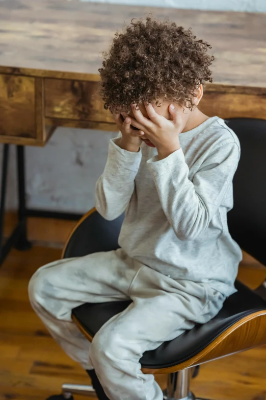 a little boy sitting on a chair with his head in his hands, pexels, wearing a track suit, she is distressed, sitting on a mocha-colored table, hair covering eyes