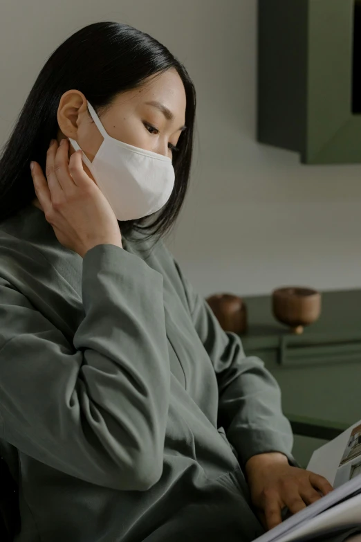 a woman wearing a face mask while reading a magazine, inspired by Li Di, trending on pexels, dressed in a green robe, grey, pondering, korean