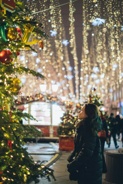 a woman standing in front of a christmas tree, happening, bright city lights, square, thumbnail, decorations