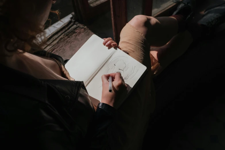 a woman is drawing on a piece of paper, a drawing, by Emma Andijewska, pexels contest winner, curled up on a book, near a window, holding notebook, single portrait