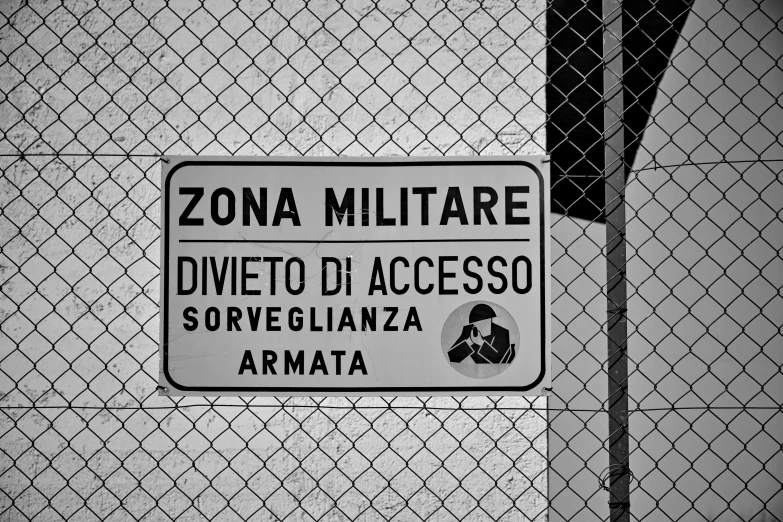 a sign is posted on a chain link fence, a poster, by Vito D'Ancona, pixabay, graffiti, military buildings, mozzarella everywhere, fotografia blanco y negro, private academy entrance