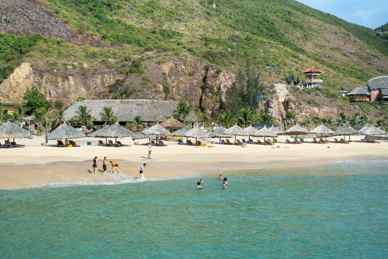 a group of people standing on top of a sandy beach, vietnam, huts, lush surroundings, piroca