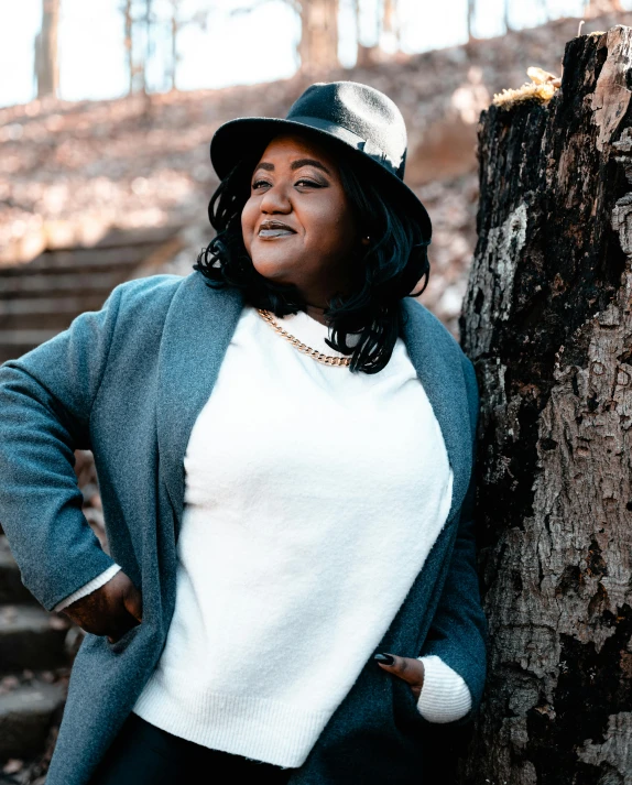 a woman standing next to a tree in a forest, an album cover, inspired by Mona Moore, trending on unsplash, happening, plus size woman, wearing a fedora, gray hoodie, bbwchan