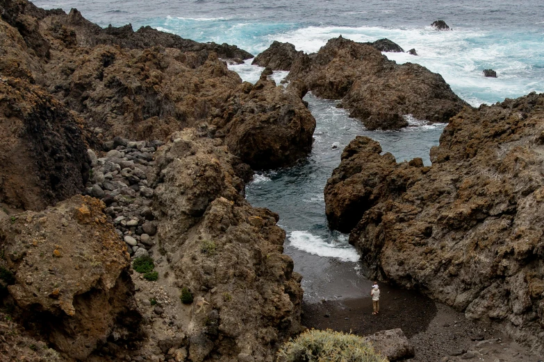 a man standing on top of a rocky beach next to the ocean, by Simon Marmion, pexels contest winner, azores, rock pools, brown, thumbnail