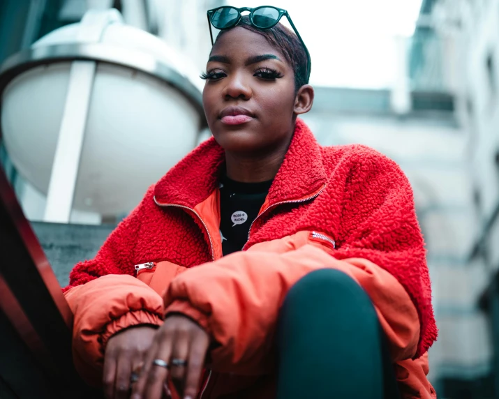 a woman sitting on the steps of a building, by Niko Henrichon, trending on pexels, orange jacket, black young woman, wearing an aviator jacket, wearing red attire