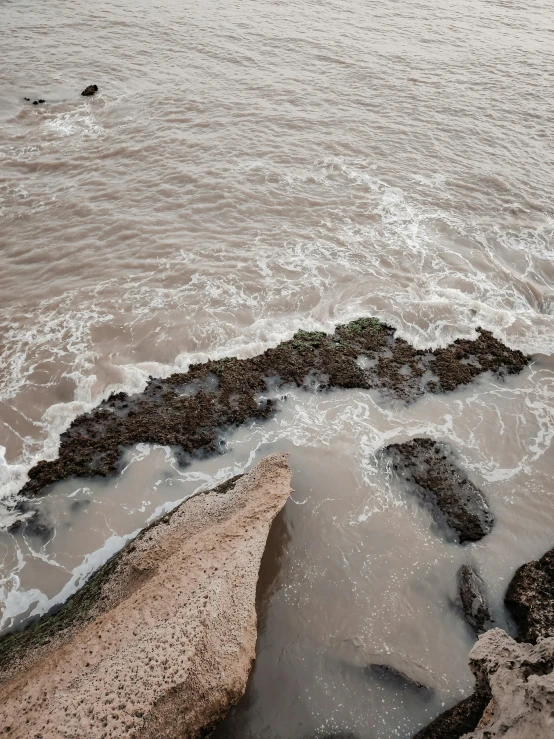 a man standing on top of a rock next to the ocean, an album cover, inspired by Andreas Gursky, unsplash, australian tonalism, brown mud, close-up from above, the thames is dry, sea spray