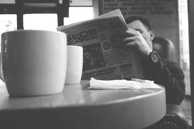 a man sitting at a table reading a newspaper, a black and white photo, by Karl Buesgen, pexels, two cups of coffee, gif, end of the day, alex yanes
