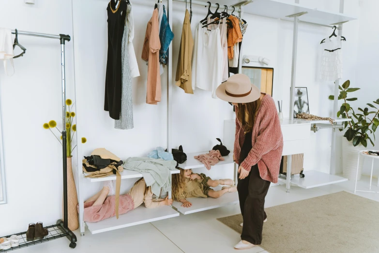 a woman standing in front of a rack of clothes, by Liza Donnelly, trending on pexels, indi creates, baggy clothing and hat, in a white room, presenting wares