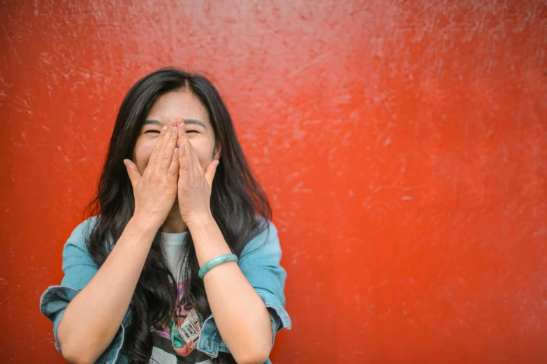 a woman covering her face with her hands, pexels contest winner, red wall, giggling, half asian, 15081959 21121991 01012000 4k
