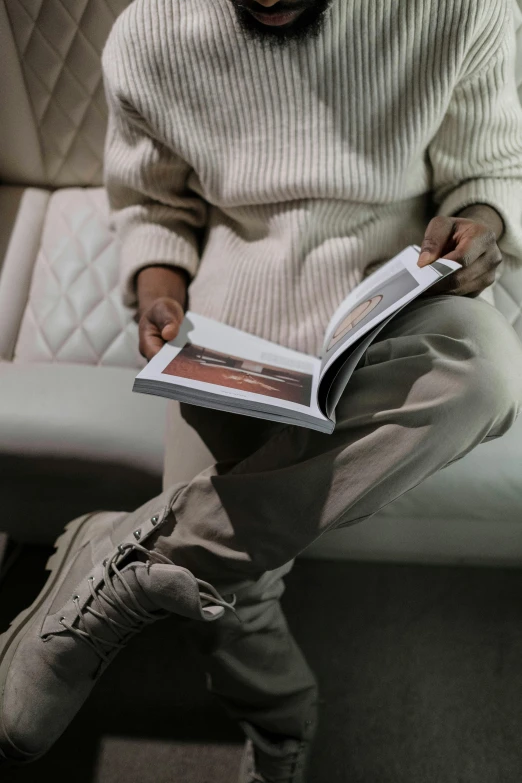 a man sitting on a couch reading a book, a picture, by Matija Jama, trending on pexels, yeezy collection, brown pants, wearing off - white style, neutral color