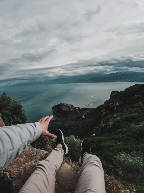 a person sitting on top of a rock next to a body of water, by Alexis Grimou, bird\'s eye view, holding his hands up to his face, overcast skies, hills and ocean