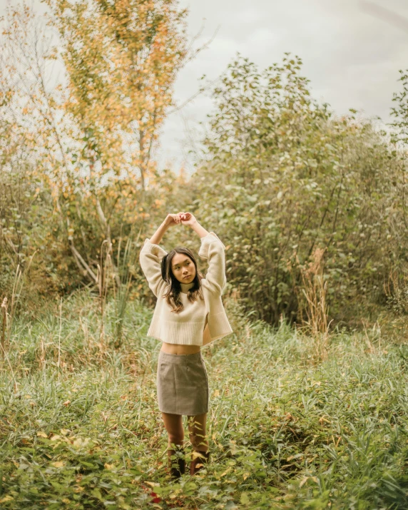 a woman standing in a field with her hands in the air, inspired by Elsa Bleda, unsplash, brown sweater, white shirt and grey skirt, non binary model, posing for a picture