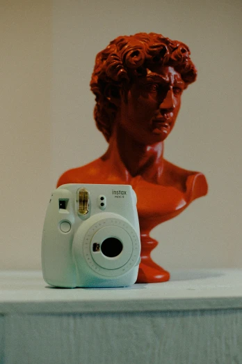 a camera sitting on top of a table next to a bust of a man, a polaroid photo, visual art, white soft leather model, vibrant colour, red mood in background, 🎨🖌️