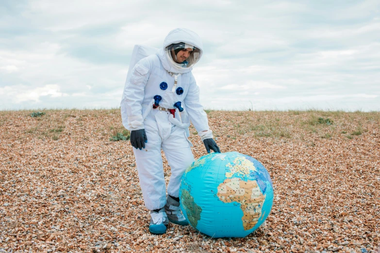 a man in a white space suit standing next to a blue globe, inspired by Storm Thorgerson, wearing dirty travelling clothes, earth outside, lunar themed attire, he is covered with barnacles