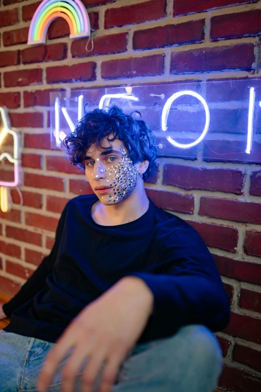 a man sitting in front of a brick wall, an album cover, by Bernie D’Andrea, trending on pexels, elaborate lights. mask on face, ethan klein, curls, an epic non - binary model