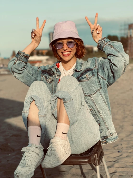 a woman sitting on top of a chair on a beach, trending on pexels, happening, an aviator jacket and jorts, peace sign, wearing a cute hat, non binary model