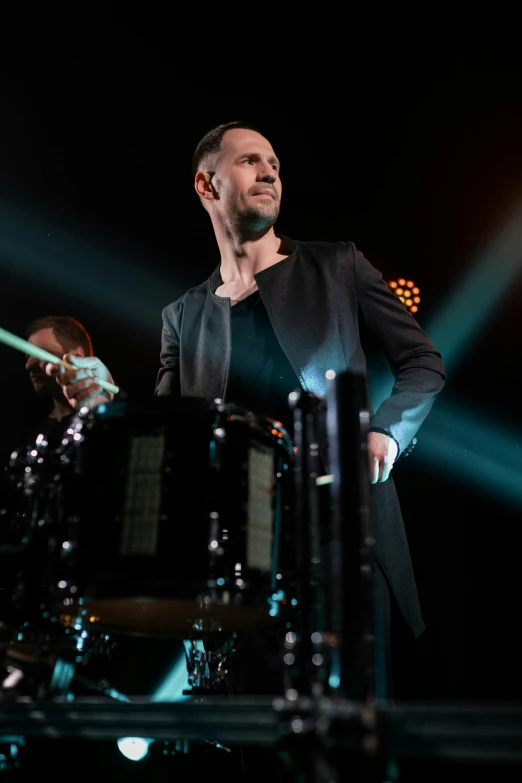 a man standing next to a drum on a stage, an album cover, inspired by Hallsteinn Sigurðsson, pexels, bauhaus, man in black suit, doug walker, flashing lights, thumbnail