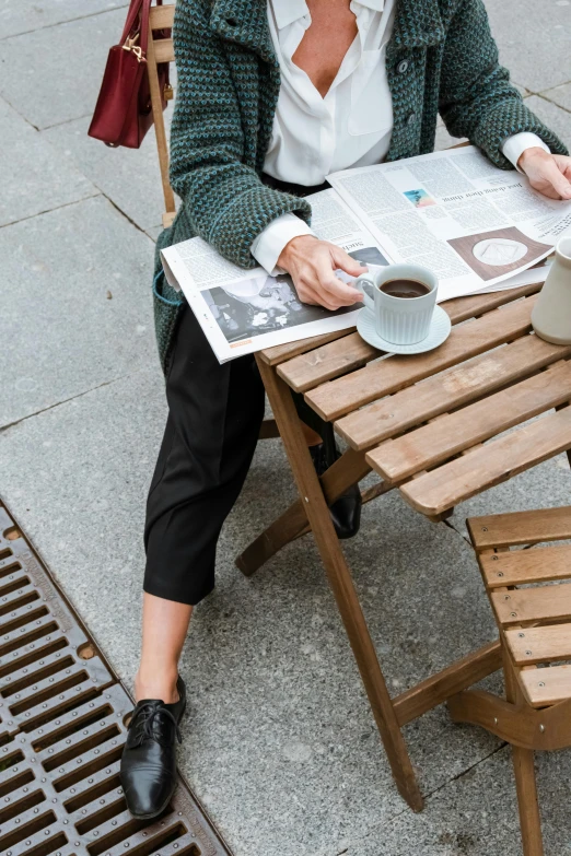 a woman sitting at a table with a cup of coffee, inspired by Ruth Orkin, happening, black loafers, brown pants, newspaper, street wear