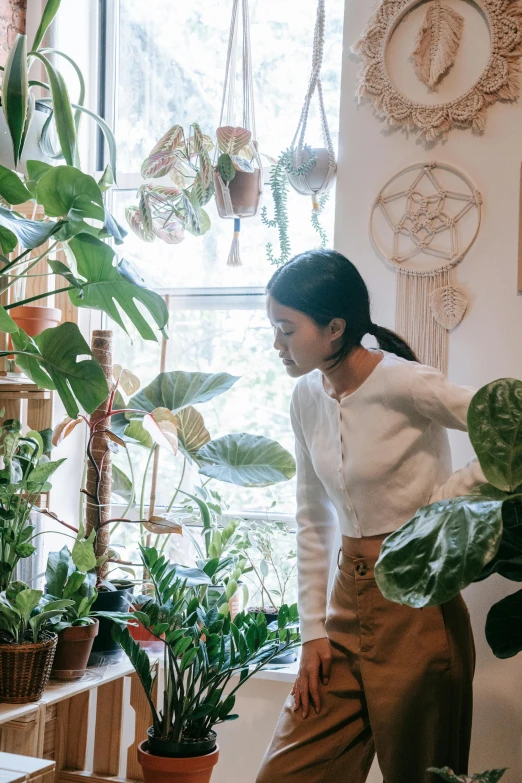 a woman standing in front of a window next to a potted plant, trending on unsplash, on a planet of lush foliage, vendors, gemma chen, home display