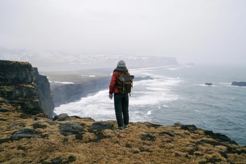 a person standing on a cliff overlooking the ocean, by Louisa Matthíasdóttir, pexels contest winner, with a backpack, overcast, rough seas in background, overlooking a valley