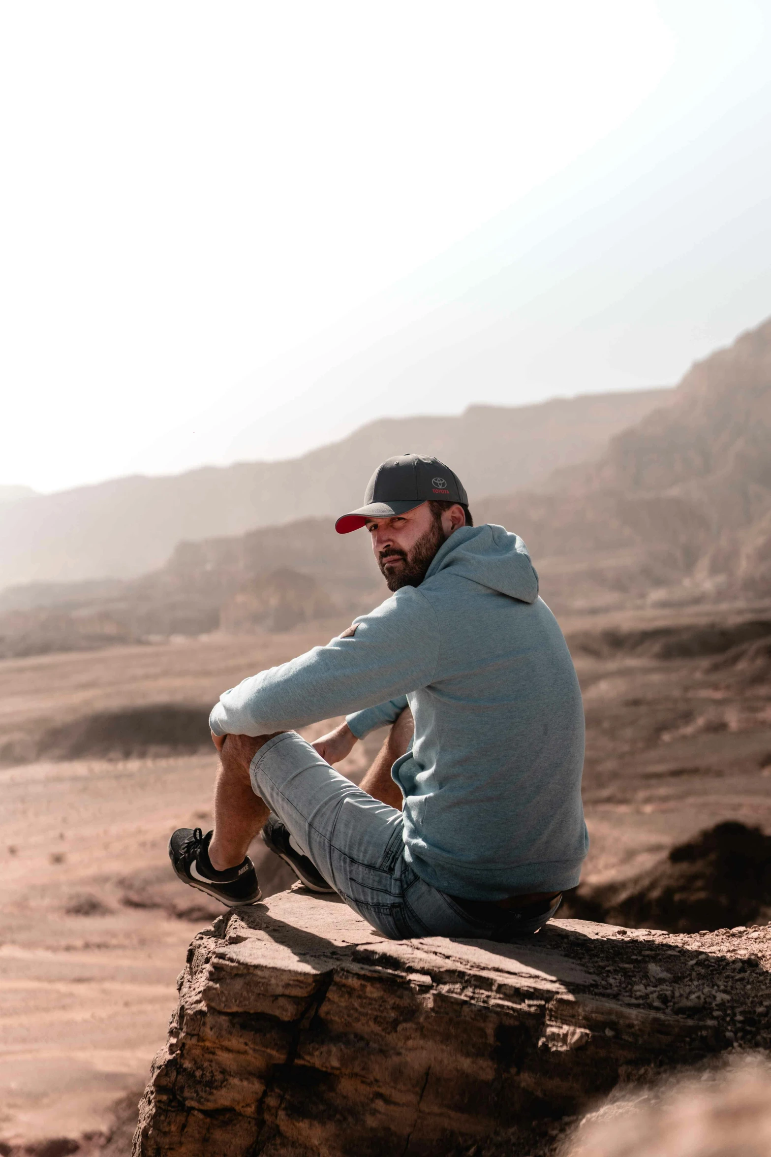 a man sitting on a rock in the desert, wearing a baseball cap backwards, eytan zana, relaxed colors, avatar image