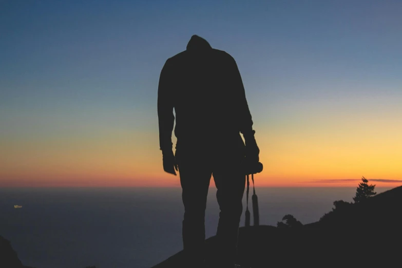 a man standing on top of a mountain holding a camera, by Niko Henrichon, pexels contest winner, romanticism, back lit, avatar image, carrying survival gear, calm night. over shoulder shot