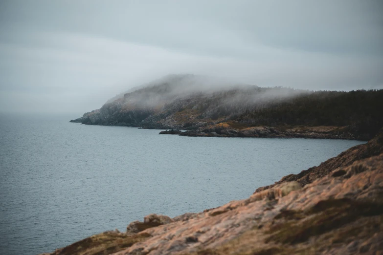 a body of water with a mountain in the background, inspired by Oluf Høst, pexels contest winner, foggy day outside, archipelago, on a cliff, faded colours