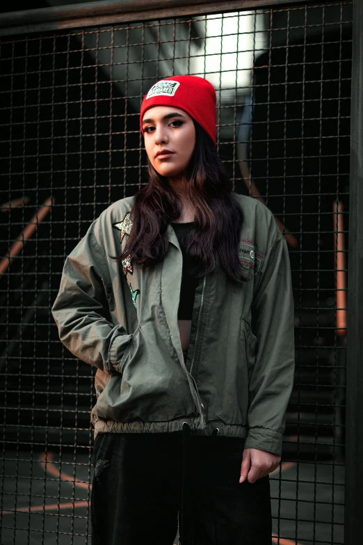 a woman standing in front of a fence wearing a red hat, a colorized photo, inspired by Elsa Bleda, trending on pexels, graffiti, wearing dark green bomber jacket, young woman with long dark hair, on a gray background, military clothing