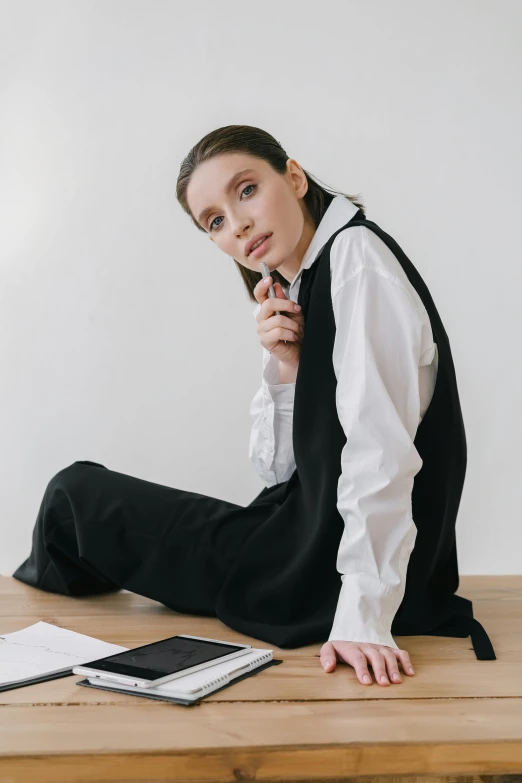 a woman sitting on top of a wooden table, inspired by Josefina Tanganelli Plana, trending on pexels, wearing school uniform, black vest, in a white room, fashion editorial