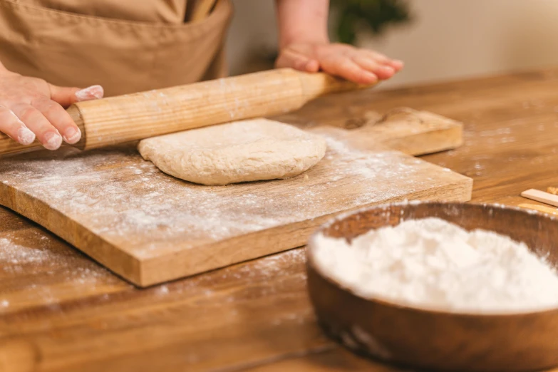 a person rolling out dough on a wooden table, by Sylvia Wishart, trending on pexels, holding a wooden staff, covered in white flour, thumbnail, petite