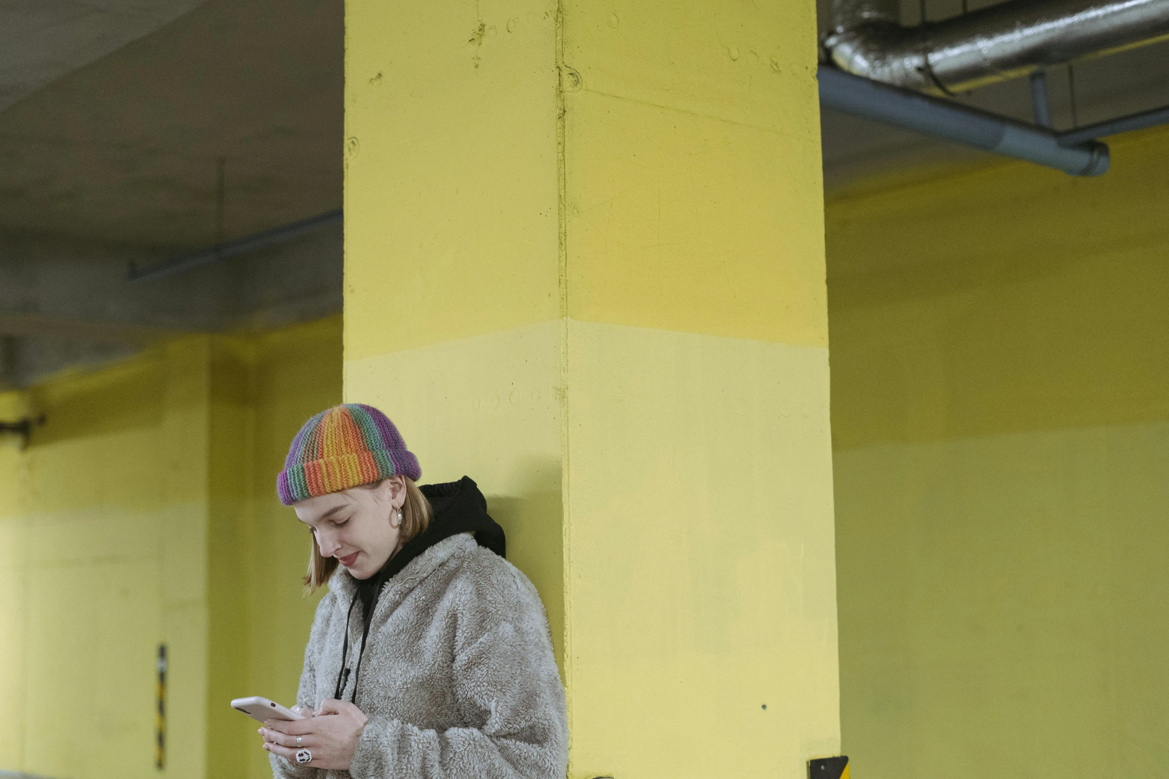 a woman standing in a parking garage looking at her cell phone, by Grytė Pintukaitė, graffiti, gradient yellow, knitted hat, production photo, square