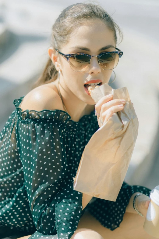 a woman sitting on the ground eating a donut, with dark brown sunglasses, holding a baguette, plastic wrap, creamy skin