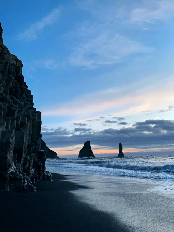 a man standing on top of a beach next to the ocean, tall stone spires, black sand, cliff side at dusk, three towers