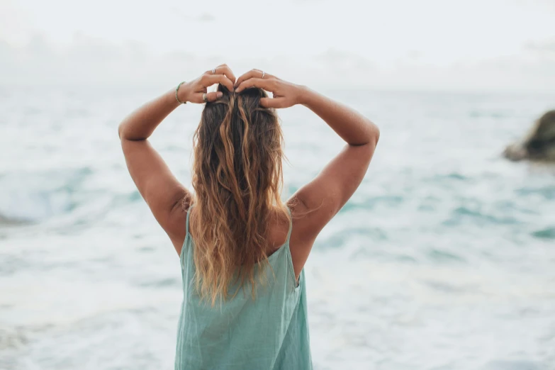 a woman standing on top of a beach next to the ocean, trending on pexels, hands in her hair. side-view, teal aesthetic, her hair is natural disheveled, arched back