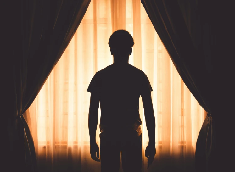 a silhouette of a man standing in front of a window, a stock photo, pexels, romanticism, glowing drapes, in my bedroom, in front of an orange background, dark skin