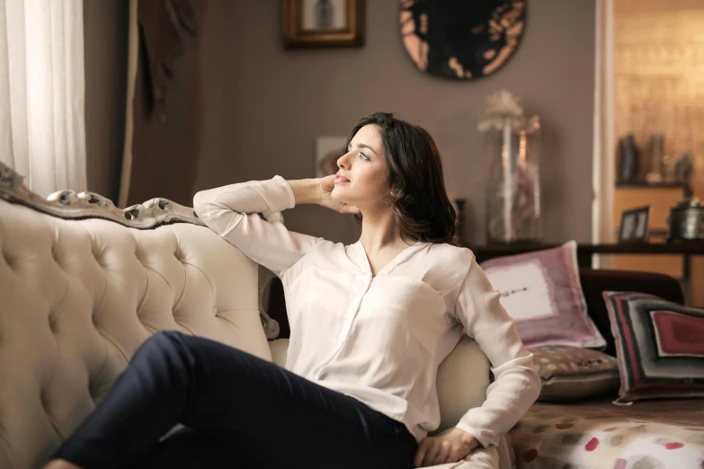 a woman sitting on a couch in a living room, inspired by Alexander Roslin, pexels contest winner, arabesque, thoughtful pose, wearing a linen shirt, elegant profile pose, lovely woman