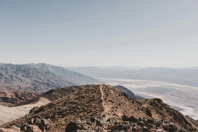 a person standing on top of a mountain, by Morgan Russell, unsplash contest winner, death valley, view from the distance, rocky ground with a dirt path, slightly tanned