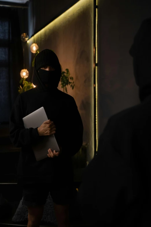a person standing in a dark room holding a book, man steal computers, black hood, holding notebook, two male