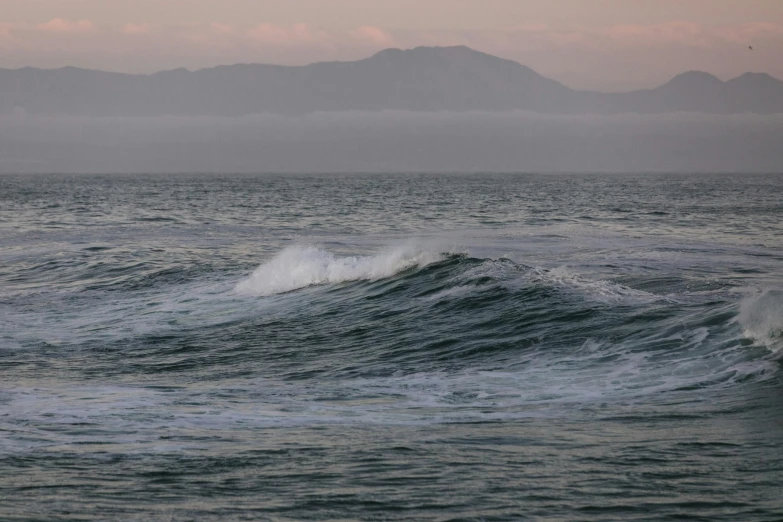 a man riding a surfboard on top of a wave in the ocean, by Charlotte Harding, pexels contest winner, romanticism, overcast dusk, mountains in distance, light pink mist, photograph of san francisco