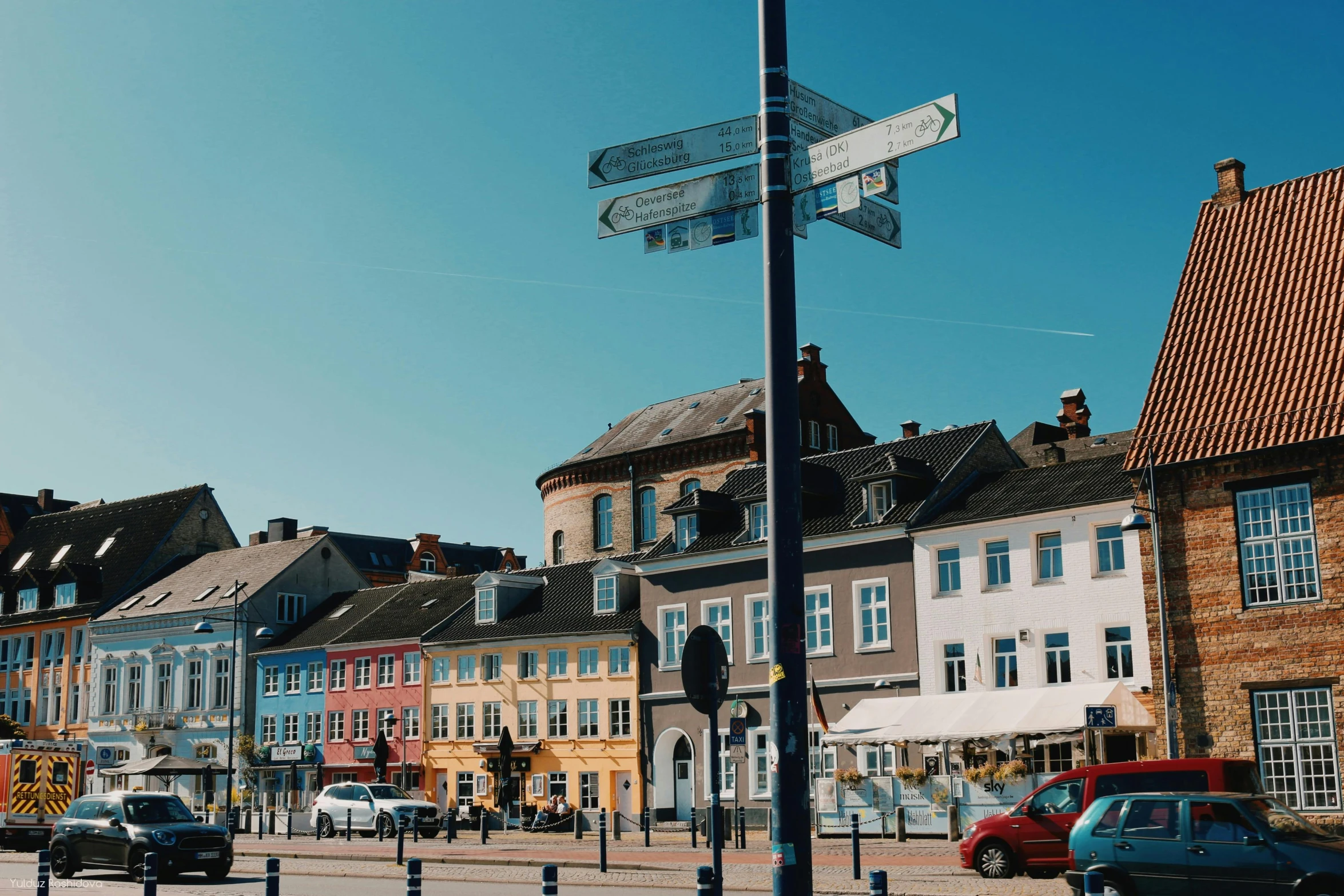 a group of cars driving down a street next to tall buildings, by Tobias Stimmer, unsplash, maryport, market square, swedish houses, in the foreground a small town