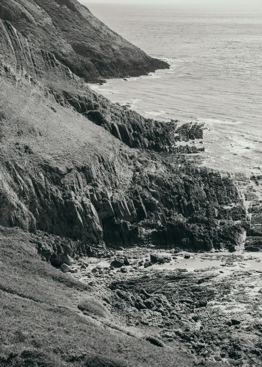 a man standing on top of a cliff next to the ocean, a black and white photo, inspired by Thomas Struth, les nabis, shiny layered geological strata, 1950s photograph, extra detail, pembrokeshire