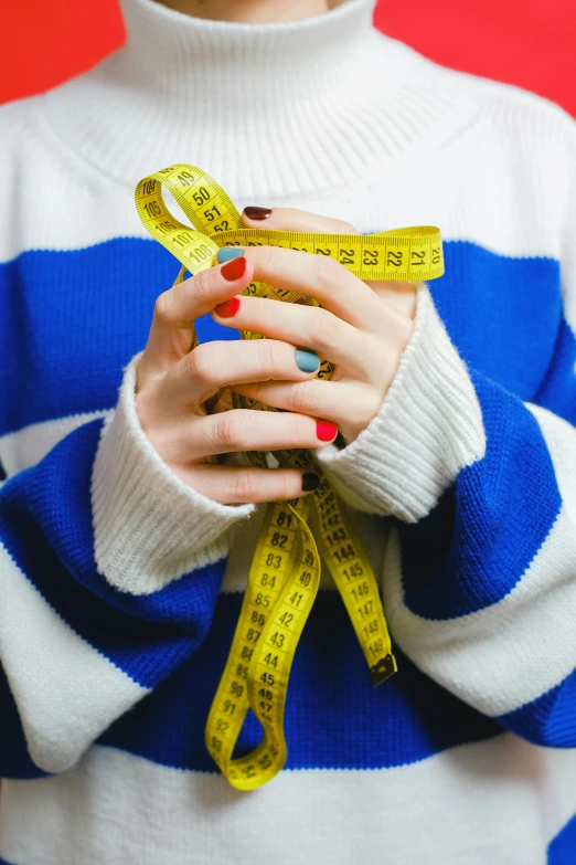 a woman holding a measuring tape in front of her face, by Julia Pishtar, colourful clothes, smallest waistline, wearing a sweater, blue and yellow color theme