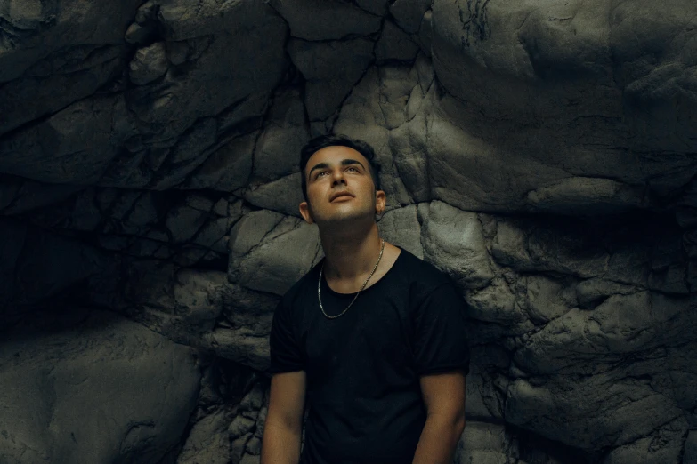 a man standing in front of a rock wall, an album cover, inspired by Alexis Grimou, unsplash contest winner, realism, dramatic lighting; 4k 8k, he is wearing a black t-shirt, kailee mandel, flume